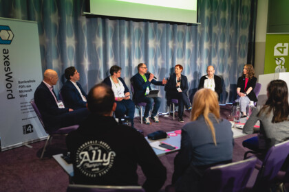 Panel-Diskussion. 7 Leute sitzen im halben Stuhlkreis nebeneinander. Einige aus dem Publikum sind von hinten abgebildet.