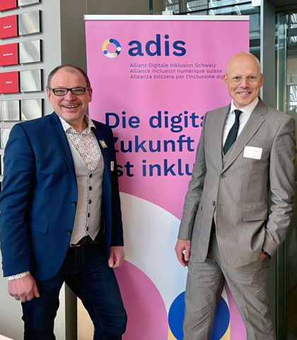 Markus Erle und Markus Riesch vor einem Banner mit der Aufschrift: "ADIS. Die digitale Zukunft ist inklusiv."